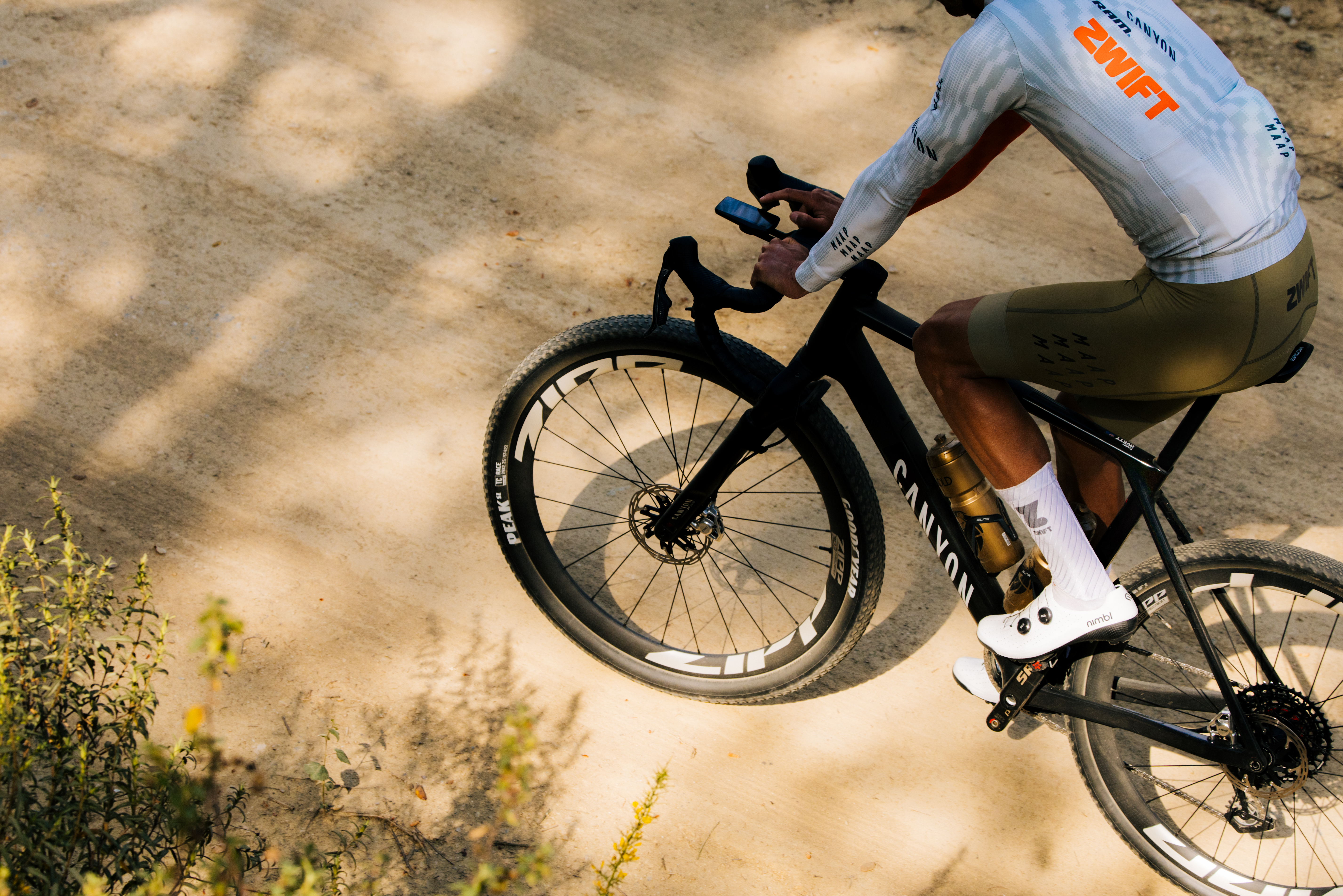 Cyclist on a Canyon bike checks a handlebar device while riding on a dirt path.